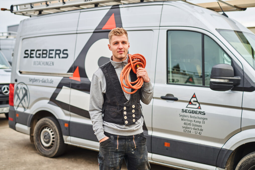 Handwerker mit Kabel vor einem Lieferwagen mit dem Logo „Segbers Bedachungen“.