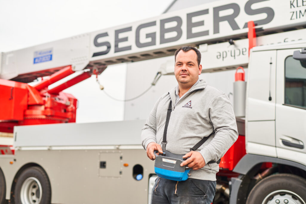 Mann in grauem Pullover mit Steuergerät in der Hand, steht vor einem großen Fahrzeug mit Hebebühne.