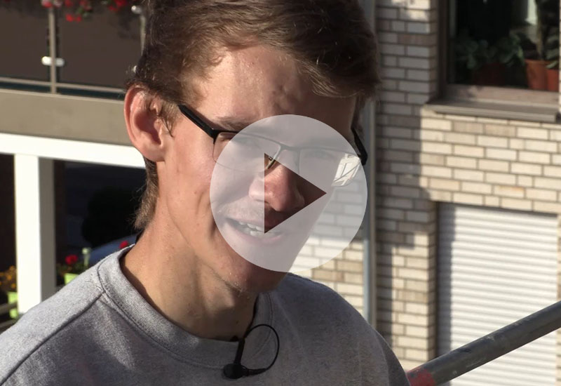 Junger Mann mit Brille und Mikrofon im Freien, halb im Schatten, mit großem Play-Symbol über dem Bild.