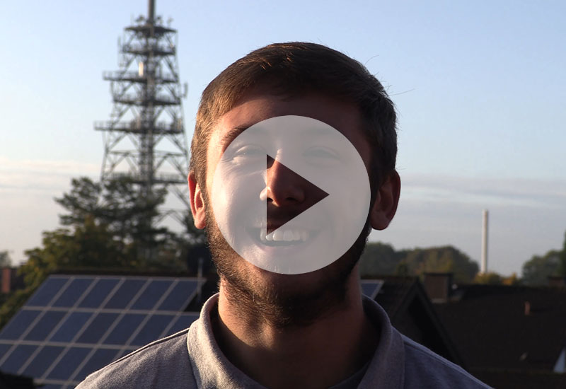 Junger Mann mit Bart lächelt vor Solarpanels und Antenne. Play-Button über seinem Gesicht.